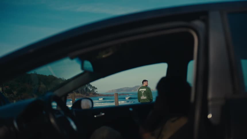 A man framed by a car window stands and contemplates the vast ocean before turning and walking towards the camera