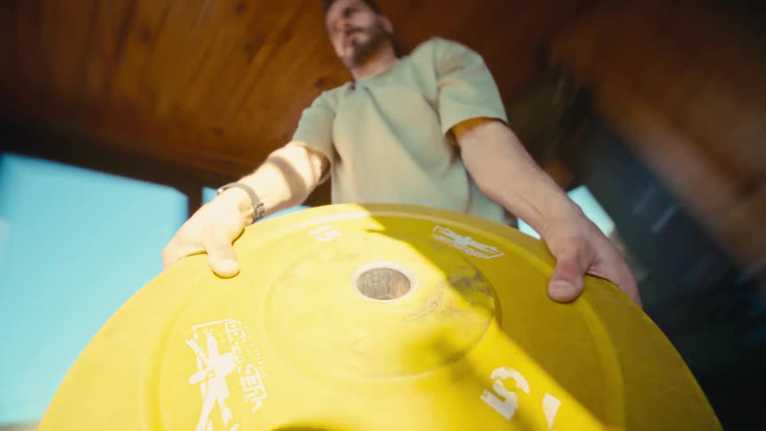 Dynamic low-angle POV of a muscular man doing an intense workout with a yellow weight plate in his home gym