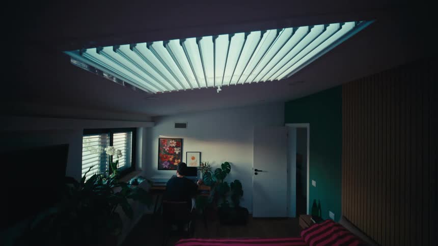 A man enters his modern bedroom with a large skylight, sits at his desk, and begins working on his laptop