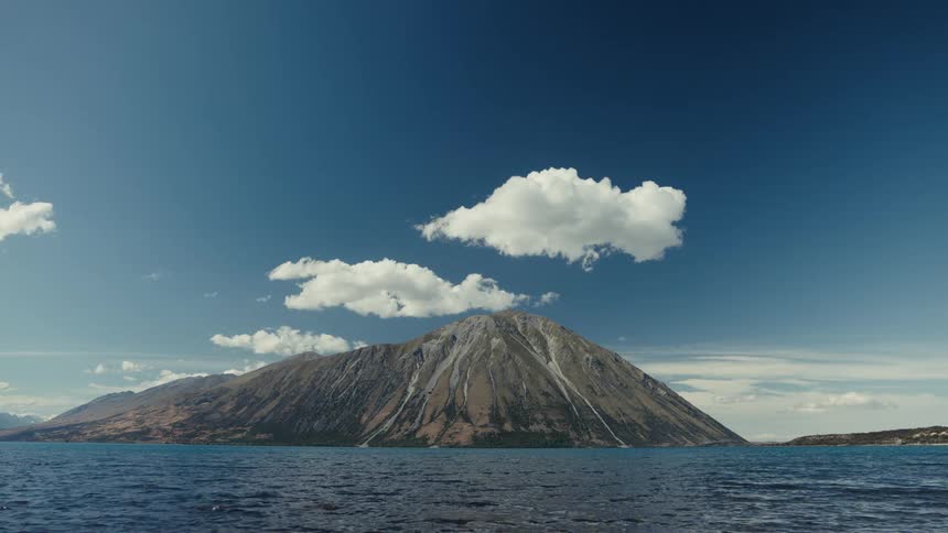 A majestic mountain sits across a vast blue lake under a clear sky with scattered clouds