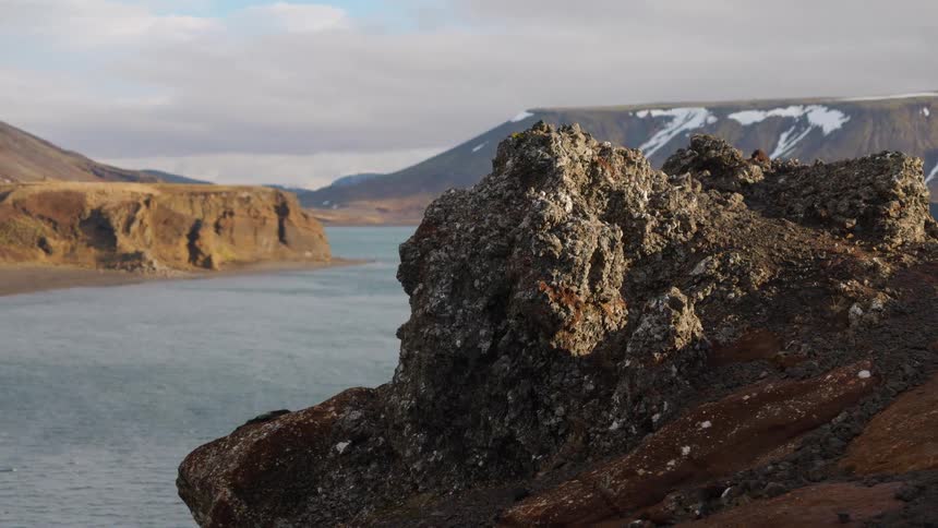 A rugged volcanic rock formation sits on a tranquil coastline, overlooking the water with snow-capped mountains in the background