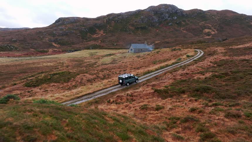 Aerial view of a green Land Rover Defender driving a remote dirt track towards an isolated stone cottage in the Scottish Highlands