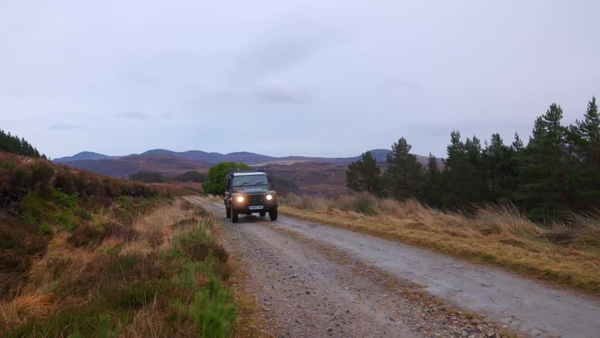 An iconic green Land Rover Defender drives along a remote, wet gravel track in the Scottish Highlands