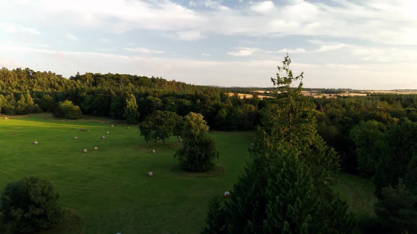 Aerial footage glides over a lush forest, revealing a tranquil meadow dotted with hay bales at golden hour