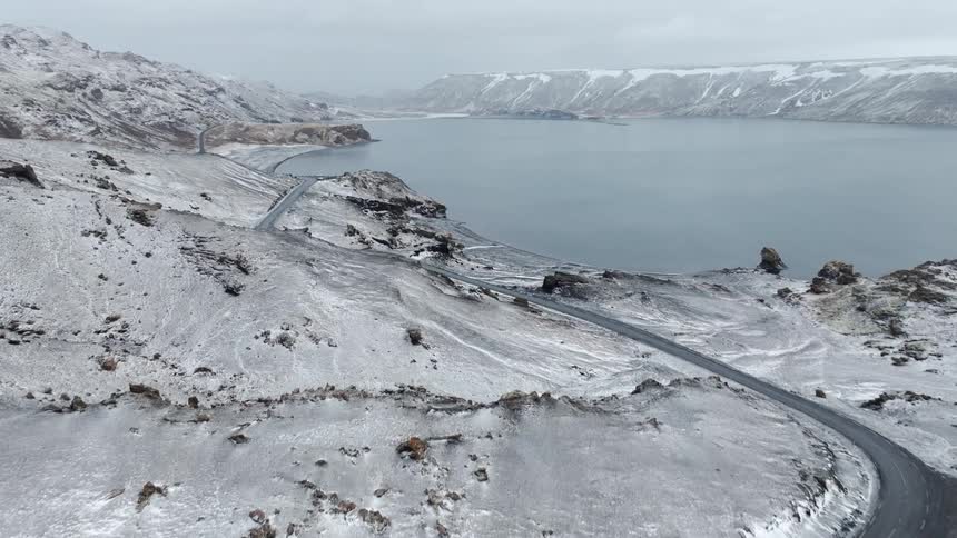 An epic aerial view follows a winding road along a vast lake through a snow-covered volcanic landscape in Iceland