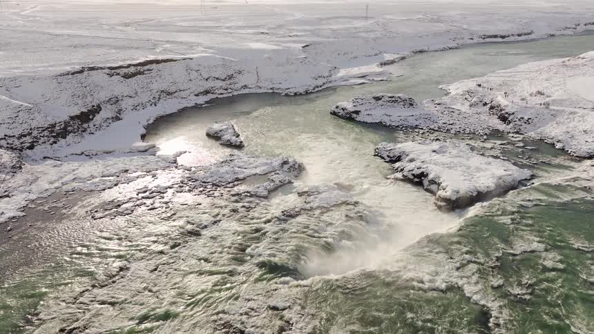 Aerial view of a powerful waterfall cascading through a vast, snow-covered landscape on a bright winter day