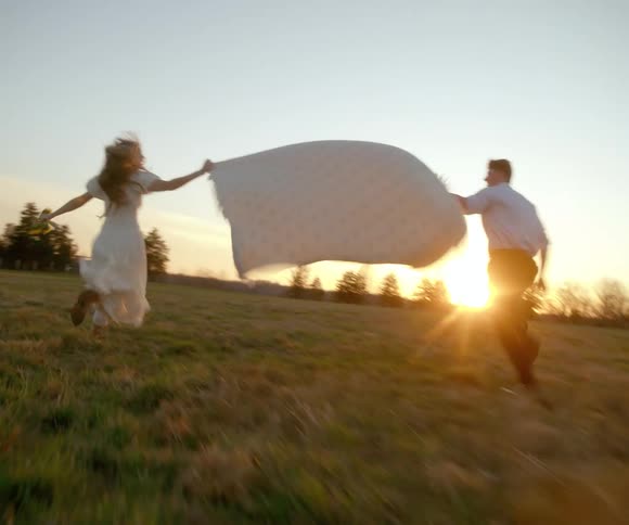 A romantic couple runs in slow motion through a golden meadow towards the sunset, holding a large billowing blanket