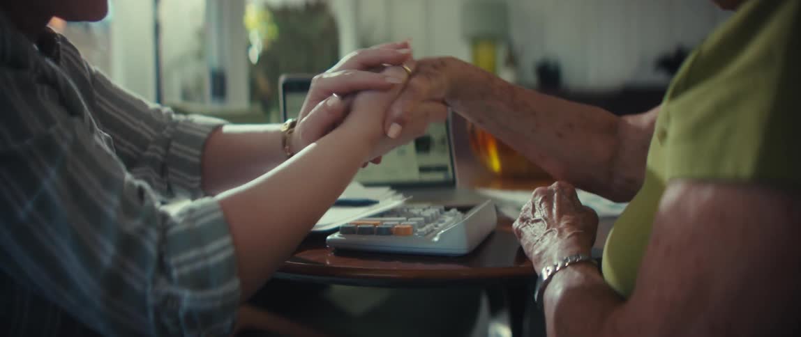 A caring financial advisor holds an elderly woman's hand reassuringly during a consultation in a brightly lit home office