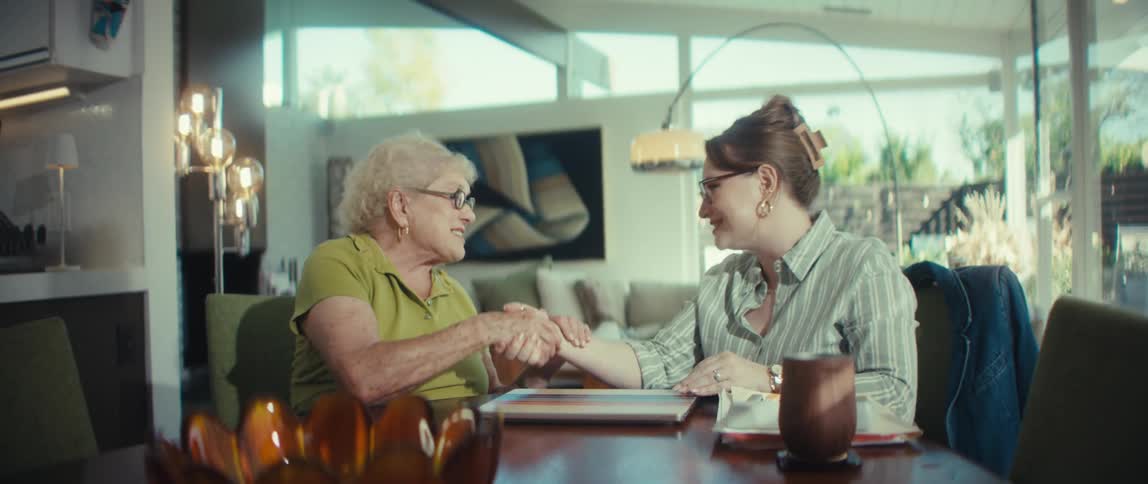 A young woman and a senior woman happily shake and hold hands across a dining table in a sunlit modern home