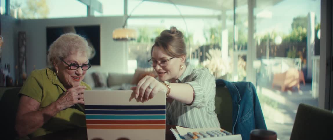 A young woman helps an elderly woman with her finances on a laptop, both smiling happily in a modern home