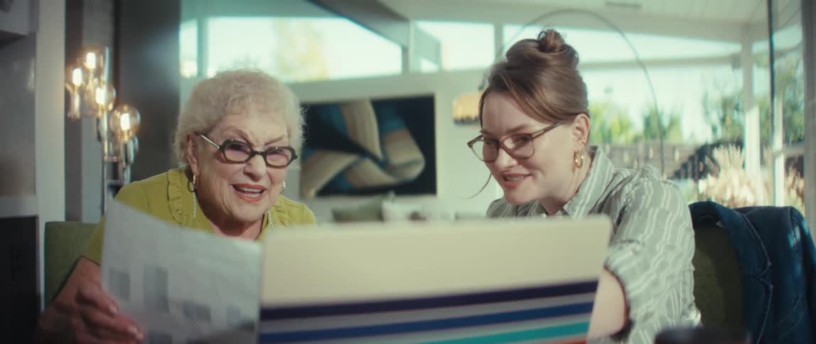 A caring young woman assists a smiling senior with her finances, looking at documents and a laptop together at home