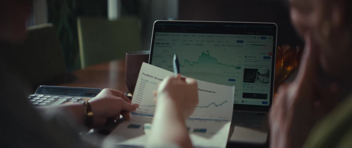 Over the shoulder view of two people discussing financial portfolio growth charts on a laptop and paper
