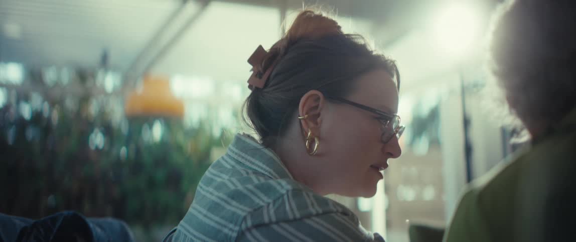 A young woman with glasses and a hair clip engages in a warm conversation with a friend, backlit by a beautiful sun flare