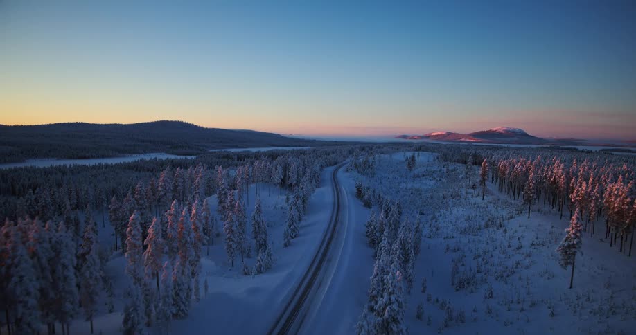 An aerial view of a winding road through a serene, snow-covered forest in Lapland as the sun sets behind distant mountains