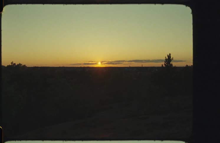A vintage film shot captures the golden sun setting over a distant city skyline, with silhouetted trees in the foreground