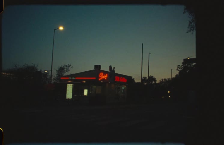 A static vintage film shot of a roadside diner with a red neon sign glowing against the dark evening sky