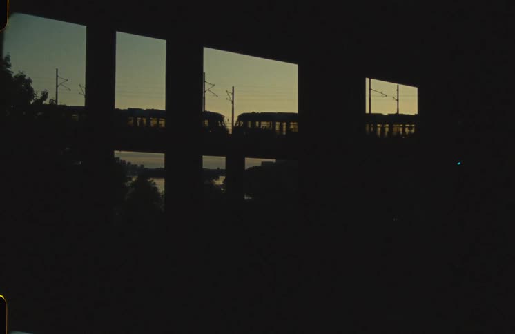 Through the panes of a dark window, two silhouetted trains pass each other on an elevated track against the tranquil twilight sky