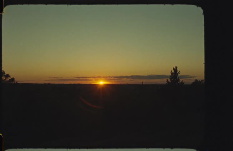 A vintage film capture shows the sun setting on the distant horizon over a silhouetted landscape