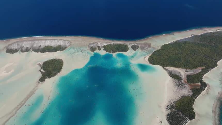 A stunning high-angle aerial view of a remote tropical atoll, where a vibrant turquoise lagoon meets the deep blue ocean, separated by white sandbars and lush green islets