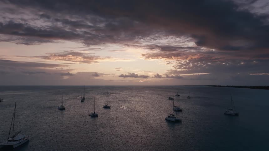 A fleet of sailboats and catamarans rest peacefully on the calm ocean during a dramatic and beautiful sunset
