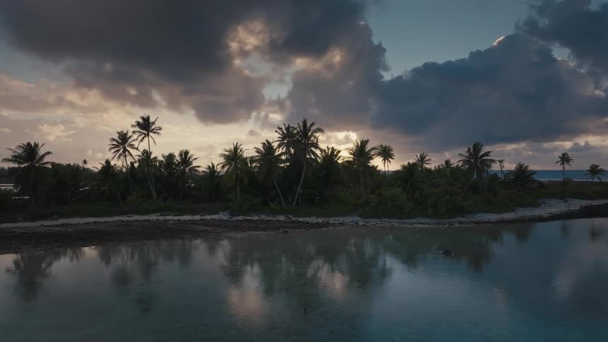 An aerial view slowly glides over calm tropical water towards a lush island with silhouetted palm trees under a dramatic cloudy sky at sunset