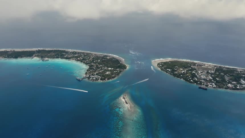 A stunning high-angle aerial view captures the beauty of a tropical atoll, showing boats moving through a pass between islands in the deep blue ocean