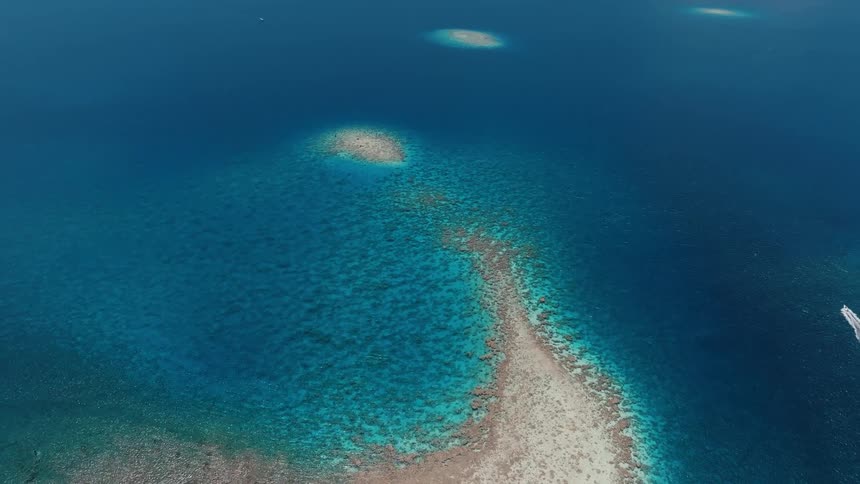 A stunning aerial view reveals a vibrant coral reef and sandbar emerging from the vast, deep blue tropical ocean as a speedboat passes by