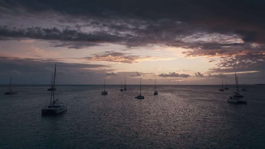 A fleet of sailboats and catamarans anchored in a calm ocean bay during a dramatic and moody sunset