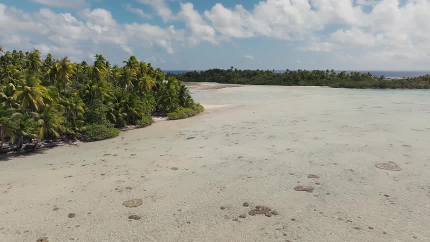 Idyllic aerial shot pulls back over a pristine shallow lagoon, showcasing a lush tropical island covered in palm trees under a partly cloudy sky