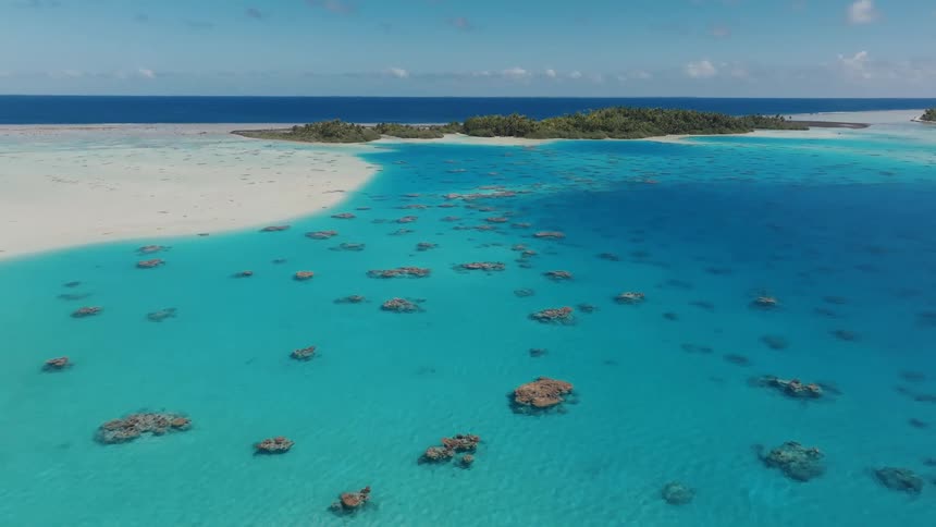 A serene aerial view shows a stunning tropical island surrounded by a vibrant turquoise lagoon with visible coral reefs and white sandbars