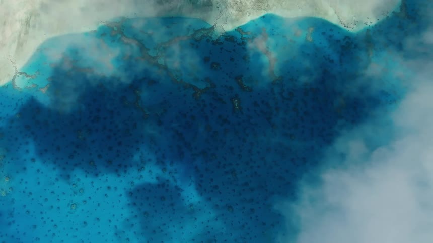 A serene aerial top-down view captures slow-moving clouds drifting over a beautiful turquoise ocean with visible coral reefs and a sandy coastline