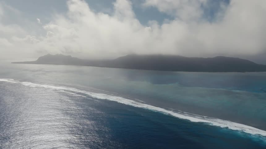 An epic aerial view shows the vast ocean with waves breaking along a reef, leading to a mysterious, cloud-covered tropical island on the horizon