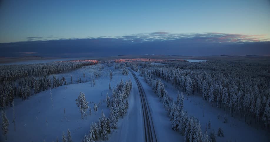 An ethereal aerial view follows a lonely railway track cutting through a vast, snow-covered forest landscape at twilight