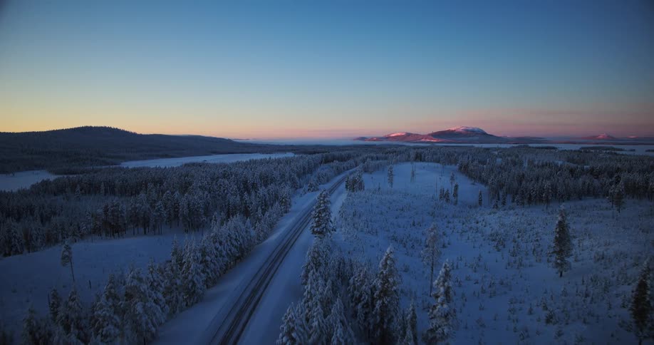 An ethereal aerial view of a snow-covered road winding through a vast winter forest landscape at twilight