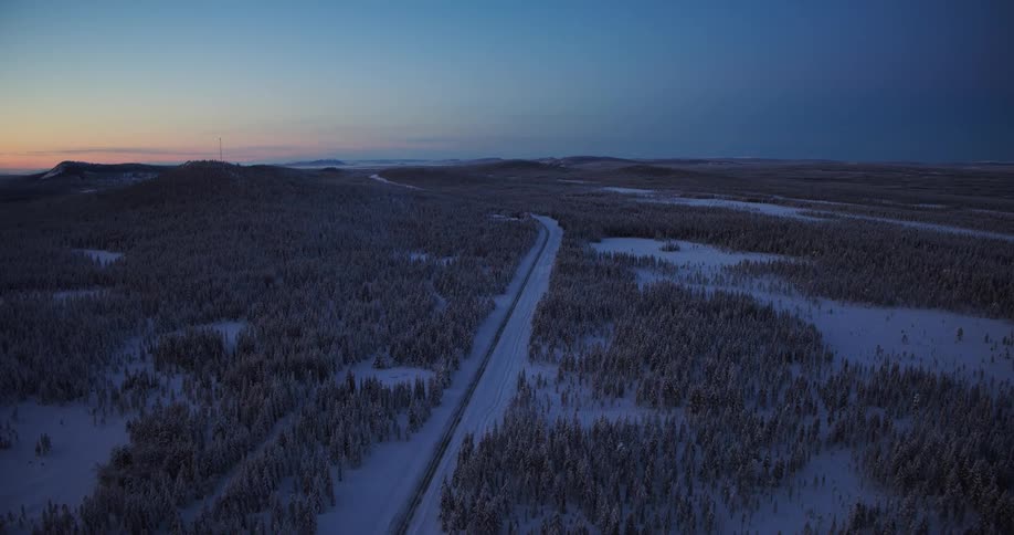 Still aerial shot of a lone road carving through an expansive snow-covered forest under a serene twilight sky in the arctic wilderness