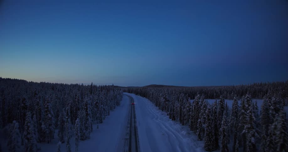 Aerial view of a lone car driving through a vast, snow-covered forest on a remote road during the tranquil blue hour