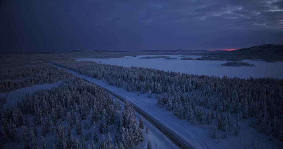 Aerial footage flies over a vast, snow-covered forest and a frozen lake during a moody blue hour at dusk in the arctic