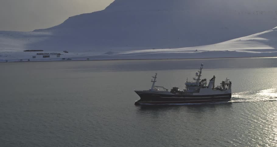 A fishing boat navigates the calm, cold waters of a fjord surrounded by a vast snowy landscape