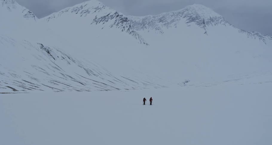 Two adventurers on an expedition trek across a vast, remote, and snowy mountain valley on a cold winter day