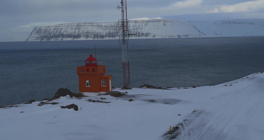 An aerial view pulls back to reveal a vibrant orange lighthouse on a remote, snow-covered cliff overlooking the sea