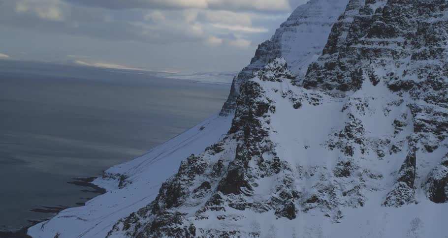 A majestic, snow-covered mountain range rises steeply from the cold ocean under a dramatic, cloudy sky