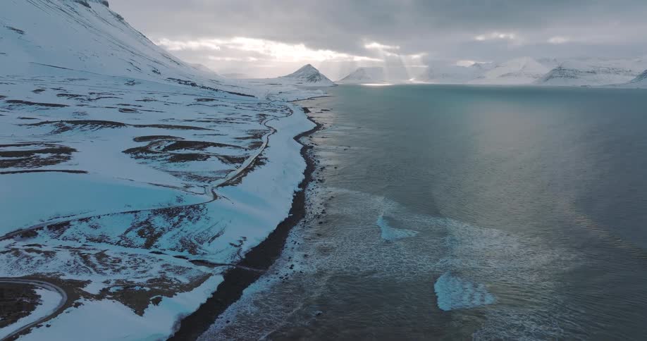 A breathtaking aerial view of Iceland's snowy coast, where ocean waves meet the winter landscape under dramatic sunlit clouds