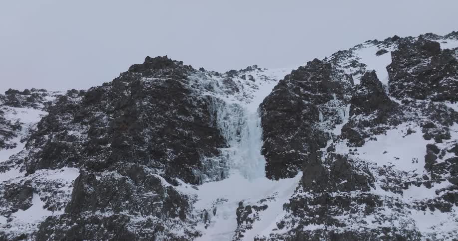 Aerial shot pulls back from a magnificent frozen waterfall cascading down a rugged, snow-covered mountain