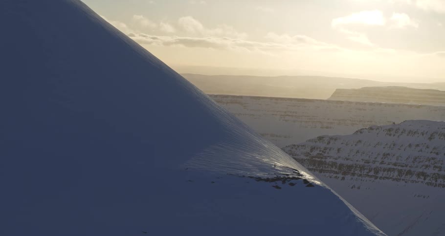 Two tiny figures hike along a vast, snowy mountain ridge during a golden sunset, with the camera slowly panning to reveal the majestic mountain range beyond