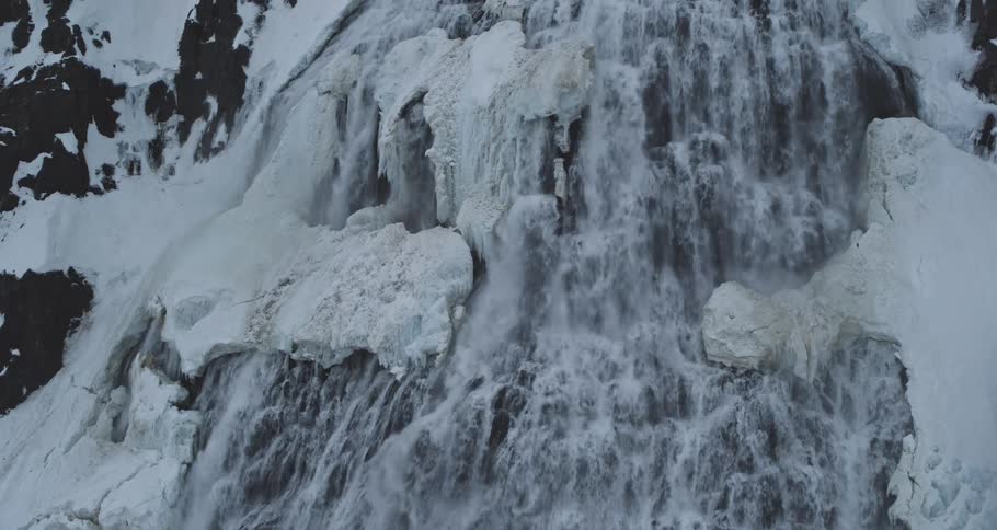 A slow upward tilt reveals a powerful, cascading waterfall rushing down a snow and ice-covered mountain in a stark winter landscape