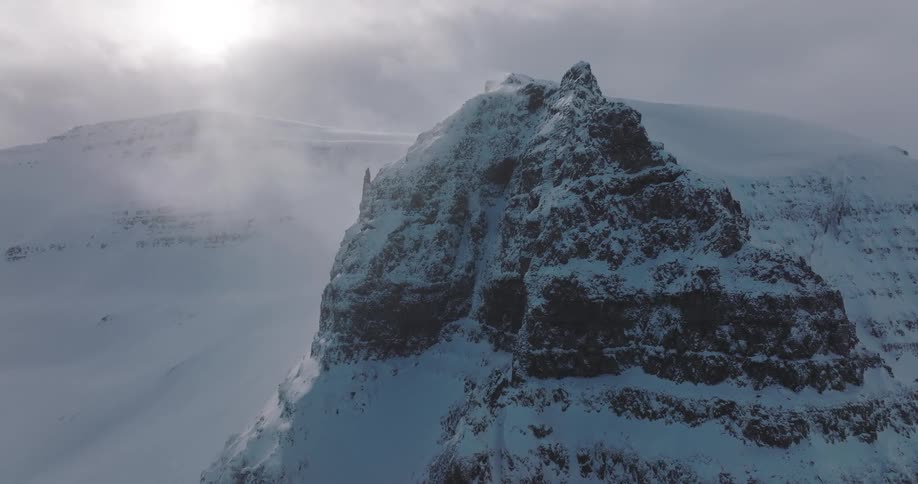A drone slowly approaches a formidable, snow-covered mountain peak as high winds create a dramatic, blizzard-like atmosphere