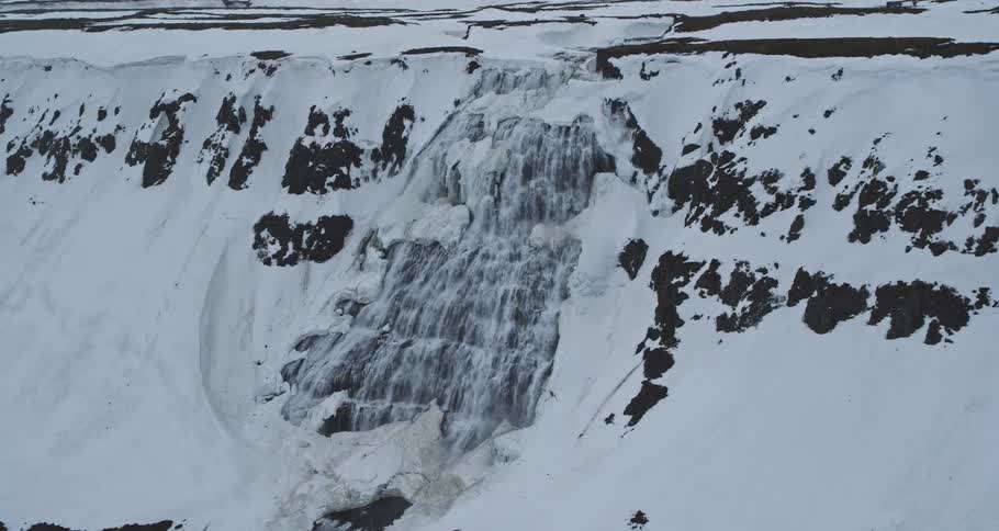 Aerial view slowly zooming in on a powerful waterfall cascading down a vast, snow-covered and partially frozen cliffside