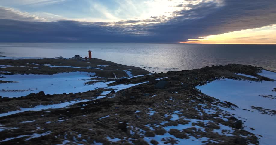 Epic aerial view of a red lighthouse on a remote, snow-covered peninsula as the sun sets over the vast, cold ocean