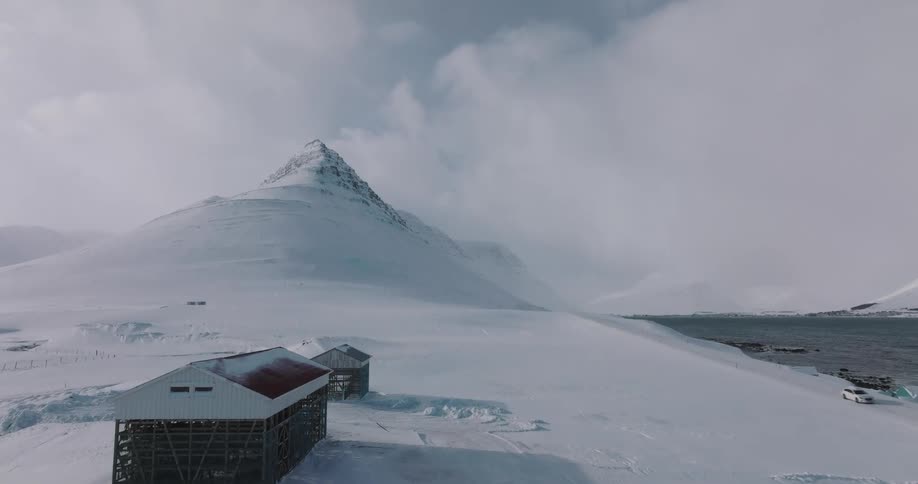 Ascending aerial shot reveals the majestic snow-covered Kirkjufell mountain and traditional fish drying racks on the Icelandic coast