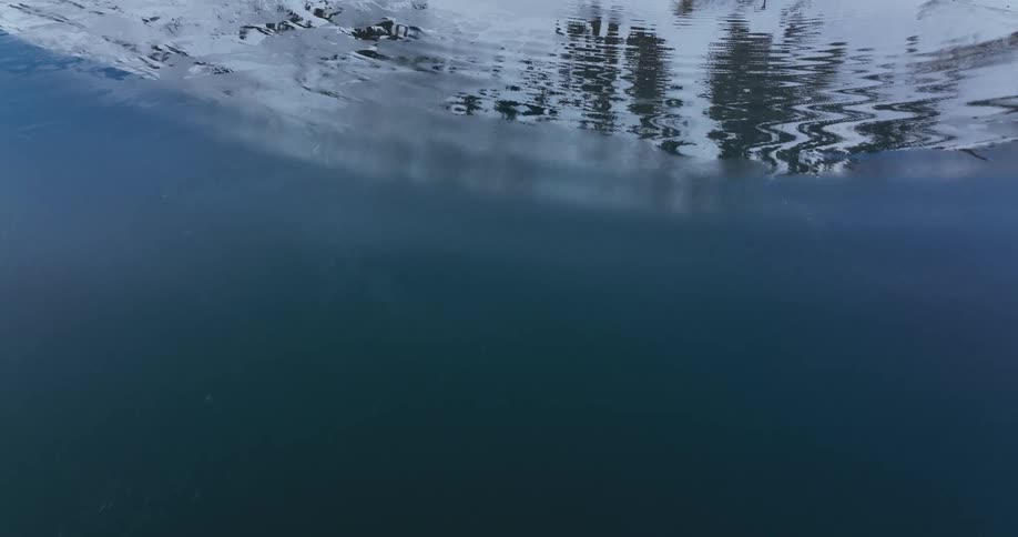 Drone pulls back from an underwater view to reveal a stunning reflection of a snowy mountain on the dark, calm water